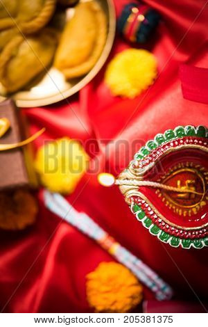 stock photo showing Diwali still life with Diya or oil Lamp, gift boxes, sweets and fire crackers with flowers arranged over red satin cloth with bokeh