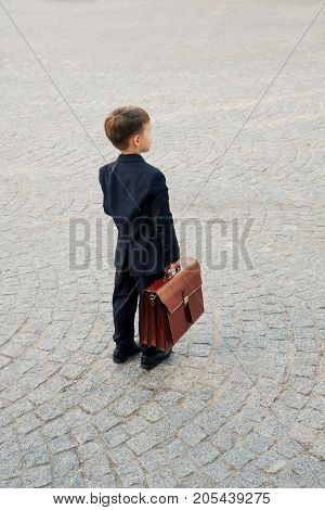 Concept future businessman. Back view of businessman in formal wear standing on pedestrian street carrying a braun briefcase looking to camera, urban gray pavage stone background. Copy space.