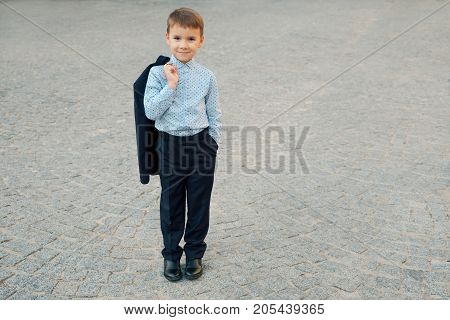Portrait elegant little man in fashionable navy blue shirt holding dark blue jacket on his back smiling and looking to camera on urban gray pavage. Young boy posing. Boy ready for school.