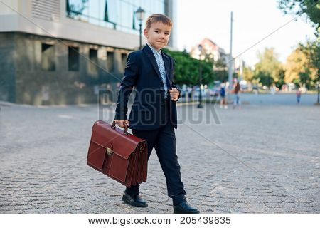 Concept future businessman. Business meeting. Low angle of future businessman in formal wear walking around the street carrying a braun briefcase and holding jacket with arm, urban background.