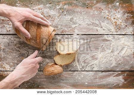 Baking background. Hands of baker cutting bread loaf with knife on rustic wooden table sprinkled with flour.