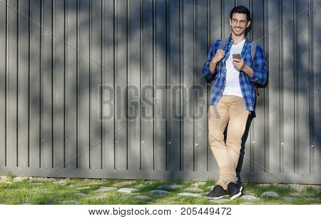 Horizontal Outdoor Portrait Of Trendy European Guy Standing Against Gray Wooden Fence Looking Leftwa