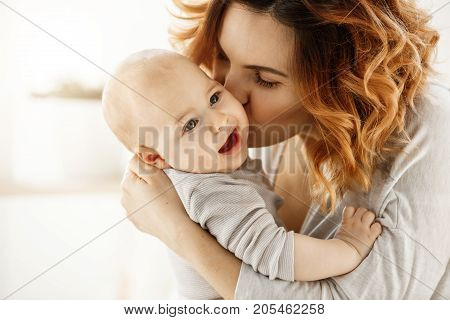 Close up portrait of young attractive mother kissing his crying baby. Kid looking aside with frightened face expression. Family concept