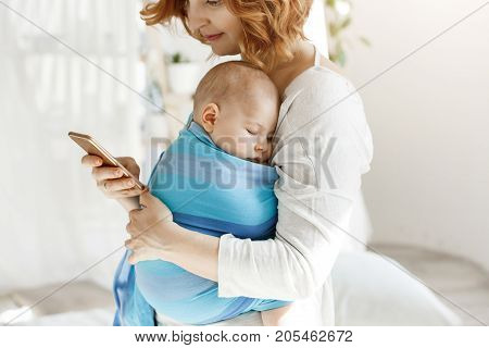 Tiny baby boy having pleasant dreams in baby slim while mother rests and looking through social networks on smartphone. Family, lifestyle concept