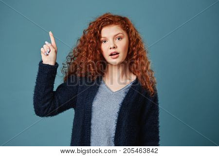 Beautiful ginger student girl with freckles in grey t-shirt and black cardigan pointing upside with happy and relaxed face expression. Copy space