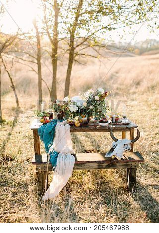 table setting, fall, travelling concept. on the wooden table for picnic standing in the field there is stunning bouquet of white flowers and cultery, decorative skull with horns placed on bench