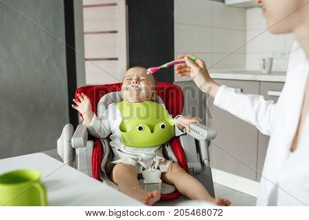 Little newborn baby sitting in baby chair with leftovers near mouth cries and refuses to eat while mother tries to feed him with a spoon