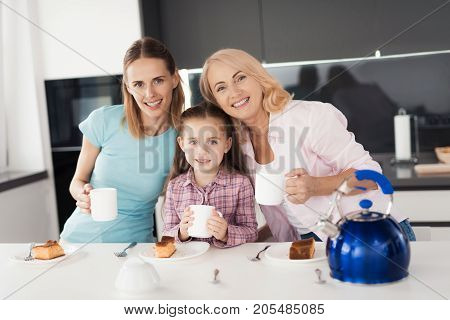 The family is drinking tea. A woman, her mother and daughter are drinking tea and posing in the kitchen. Before them are plates with a homemade pie, in their hands they hold cups of tea
