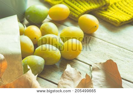 Ripe Yellow Transparent Plums in Brown Craft Paper Bag Scattered on Wood Table Dry Autumn Leaves Knitted Sweater Soft Sunlight Cozy Fall Atmosphere Toned