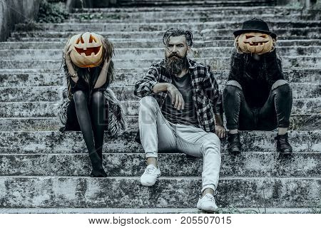 Halloween women with pumpkin heads. Friends on stony stairs. Holiday celebration concept. Halloween people and jack o lanterns. Bearded man sitting with girls.