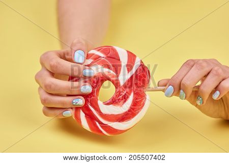 Female manicured hands with lollipop. Woman hands with beautiful manicure holding large colorful lollipop on yellow background.
