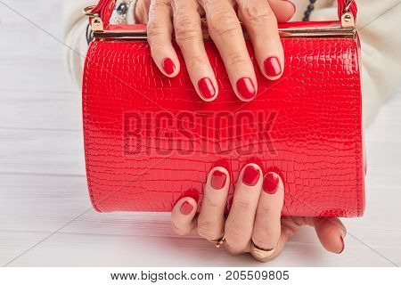 Elegant red handbag in female hands. Elderly woman hands with red manicure holding lacquered handbag close up. Womans modern accessory.