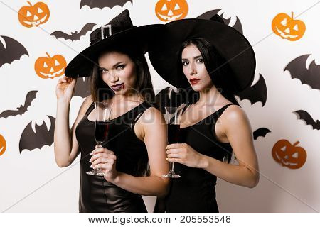 Two sexy witches are posing against a background of paper bats, dressed in black dresses and witches' hats, holding glasses of wine