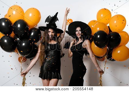 Two models in sexy black dresses posing on a white background with the clues of ballons in their hands. They are dressed in black sexy dresses and witches' hats and smile