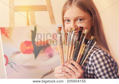 Real artist. Radiant teenage lady sitting at an easel and holding a bunch of messy painting brushes while smiling and posing for the camera.