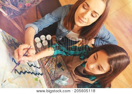 Conveying personal knowledge. Top view of a cheerful young lady and a focused teenage girl holding a painting brush together and sweeping along an art canvas during a painting session.