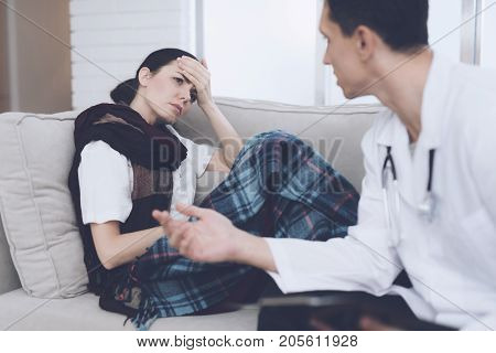 The woman fell ill and called the doctor at home. She sits on the sofa with a blanket. The doctor in a white robe is sitting next to and interrogates a woman