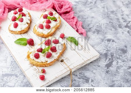 Three delicious toasts with Ricotta cheese and raspberries decorated with mint leaves on white vintage cutting board on light background
