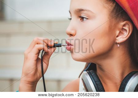 young girl puts the plug into her mouth. child in a red hat, white blouse and with grey headphones on the background of stairs