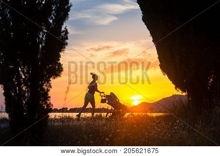 Running mother with child in stroller enjoying motherhood at sunset and mountains landscape. Jogging or power walking woman with pram on a beach at sunset. Beautiful inspirational mountains landscape.