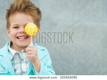 Fashion baby boy in mint jacket and sneakers stands on a gray wall background. Trendy boy with lollipop standing on the street.
