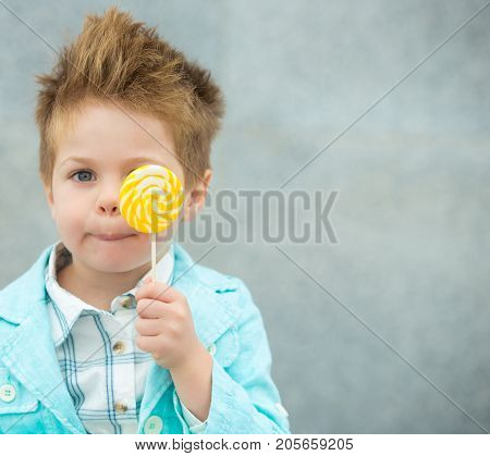 Fashion baby boy in mint jacket and sneakers stands on a gray wall background. Trendy boy with lollipop standing on the street.