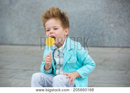 Fashion baby boy in mint jacket and sneakers stands on a gray wall background. Trendy boy with lollipop standing on the street.