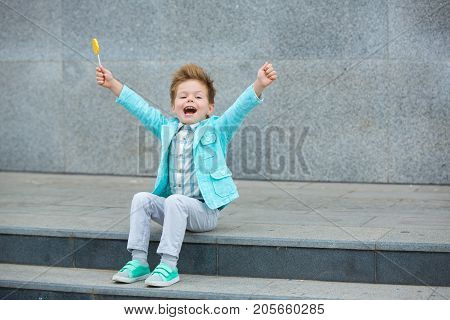Fashion baby boy in mint jacket and sneakers stands on a gray wall background. Trendy boy with lollipop standing on the street.