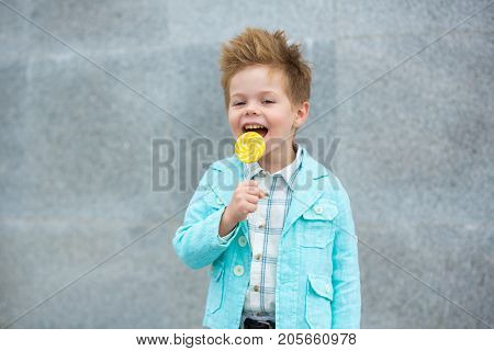 Fashion baby boy in mint jacket and sneakers stands on a gray wall background. Trendy boy with lollipop standing on the street.