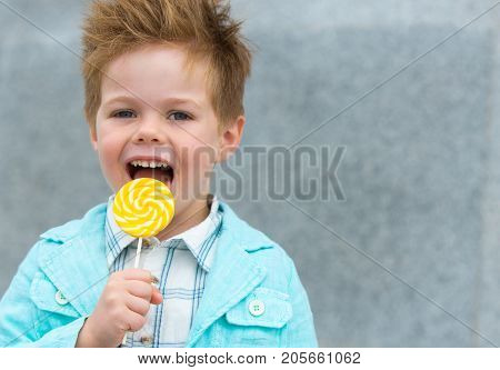 Fashion baby boy in mint jacket and sneakers stands on a gray wall background. Trendy boy with lollipop standing on the street.