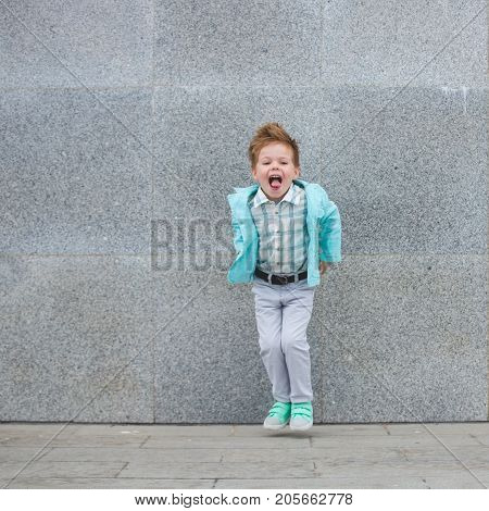 Fashion baby boy in mint jacket and sneakers posing on a gray wall background. Trendy boy standing on the street.