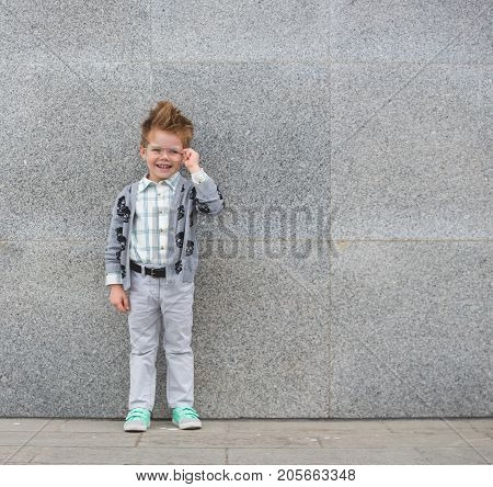 Fashion baby boy in gray jacket and mint sneakers stands on a gray wall background. Trendy boy with glasses standing on the street.