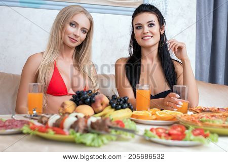 Two pretty girls in bicinis sitting at table with fruit, shashlik and drinks in sauna talking and laughing together