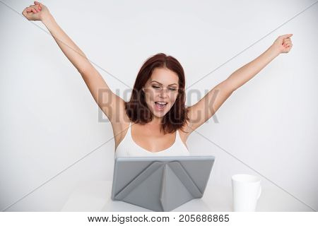 Portrait of successful young Caucasian woman sitting at digital tablet and rejoicing raising arms in triumph