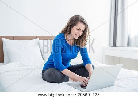 Closeup portrait of smiling middle-aged beautiful woman working on laptop and sitting with her legs crossed on bed at home