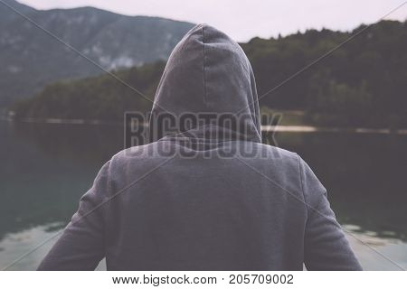 Gloomy nostalgic portrait of sad lonely melancholic adult female with hooded jacket standing on the lake shore in misty overcast morning and thinking in solitude about personal problems retro toned