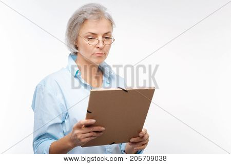 Immersed into work. Charming young woman in spectacles holding a paper folder in her hands and looking through documents in it