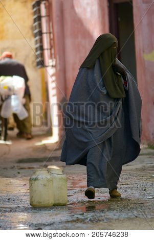woman in a black burqa goes down the street the old city cans with water nearby
