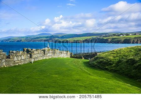 Great wall of Peel Castle constructed by vikings at Peel hill covered with green grass and beautiful coastline of Isle of Man in background