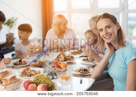 A woman in a blue T-shirt is posing against the background of a family that sits at a festive table for Thanksgiving. The family eats