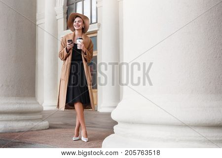 Girl in a brown coat a brown hat is walking and posing in the city interiors. The girl is smiling, checking her smartphone and drinking coffee from take away cafe. The photo looks very positive cause of the sunny and warm weather. The photo was taken in e