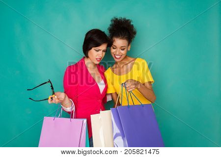 Happy multiethnic girlfriends with shopping bags. Two excited shopaholics boasting with purchases at turquoise sudio background with copy space