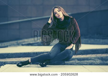 Happy young woman sitting on the sidewalk in city street. Stylish fashion model in leather fringe suede jacket and blue jeans
