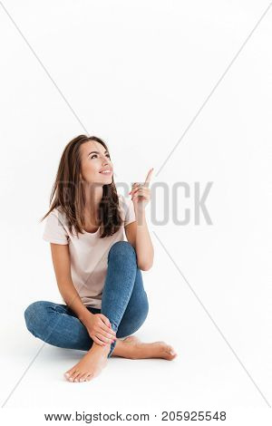 Vertical image of happy brunette woman sitting on the floor and pointing up over white background