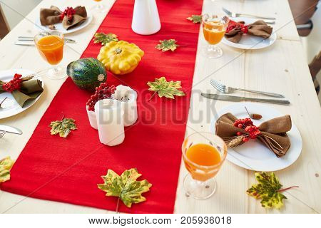 Glasses with juice, silverwear, candles and cucurbits on festive table decorated with green leaves