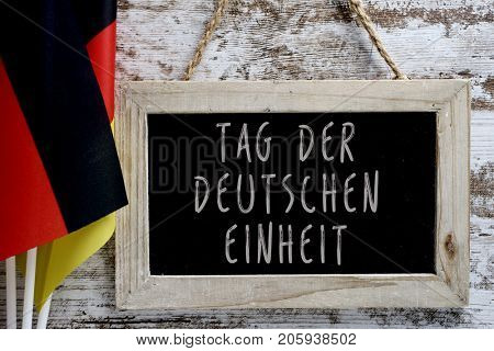 closeup of a chalkboard with the text Tag der Deutschen Einheit, Day of German Unity written in German, and some flags of Germany against a rustic wooden background