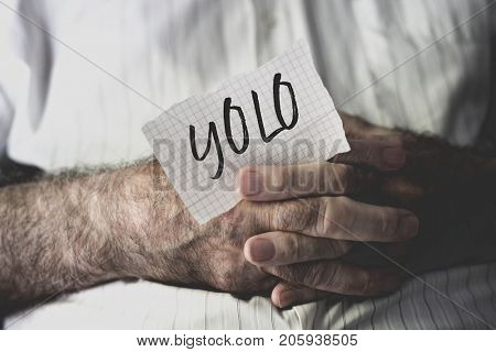 closeup of an old caucasian man with a note in his hands with the word yolo, for you only live once, written in it
