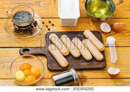Picture of ingredients for dessert, savoyardi cookies on cutting board, coffee, cream cheese, yolks, proteins