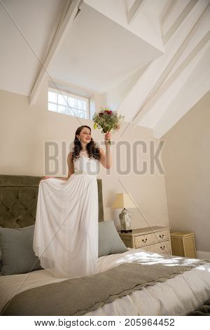 Beautiful bride holding bouquet while standing on bed at home