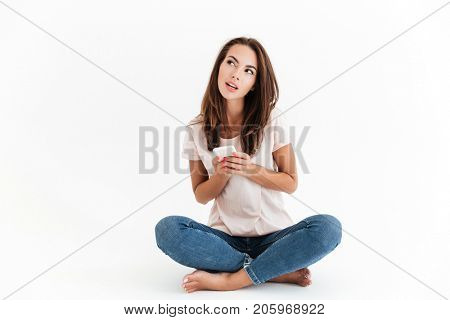 Pensive brunette woman using smartphone and looking up while sitting on the floor over white background